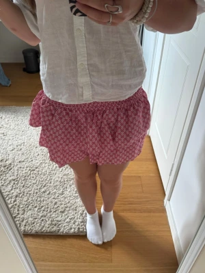 Rosa volangkjol med mönster - Söt rosa kortkjol med vita små blommo från Meet me there. Kjolen har en bred, smockad midja som ger en bekväm och flexibel passform. Perfekt för sommarens alla tillfällen och lätt att matcha med olika toppar.