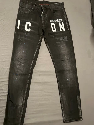 Svarta jeans - Säljer ett par svarta jeans med stor vit 'ICON'-text framtill och slitna detaljer. Jeansen har fem fickor, blå DSQ-lapp bak och är dekorerade med små färgstänk. Tillverkade i stretchig denim för en tajt passform.