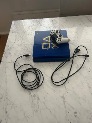 Blå PlayStation 4 Limited Edition - Säljer en blå PlayStation 4 Limited Edition med guldfärgade PlayStation-symboler på ovansidan. Konsolen kommer med en vit DualShock 4-handkontroll och tillhörande kablar. Perfekt för dig som vill ha en snygg och unik spelkonsol till samlingen.