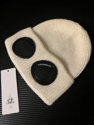Vit mössa med goggles från C.P. Company - Säljer en vit stickad mössa från C.P. Company med de ikoniska svarta gogglesen framtill. Mössan har ribbstickad struktur och uppvikt kant. Perfekt accessoar för dig som vill sticka ut och hålla dig varm under kalla dagar.