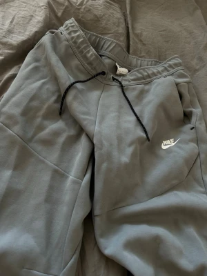 Grå Nike tech byxor med vit Nike logga - Grå Nike tech byxor med vit logga i nytt skick. Hela settets nypris ligger på 2500 kr. Byxorna: 1250 kr.