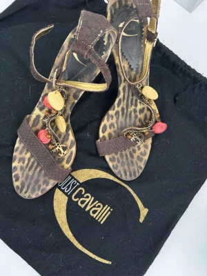 Bruna sandaler med leopardmönster Just Cavalli - Unika sandaler från Just Cavalli i brunt skinn med leopardmönstrad sula. Dekorerade med kedjor, berlocker och färgglada detaljer i guld, rött och träfärgat. Remmar runt ankeln och över foten ger en cool och trendig look.