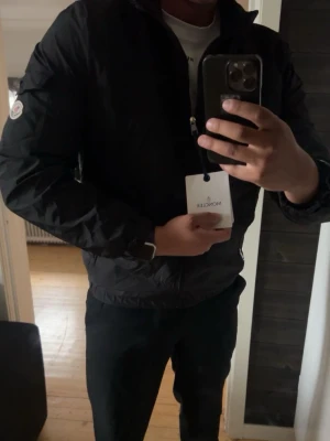 Svart vindjacka från Moncler - Snygg svart vindjacka från Moncler med dragkedja framtill och huva. Jackan har Moncler-logga på ärmen och är tillverkad i ett lätt, vindtätt material. Perfekt för dig som vill ha en stilren och funktionell jacka. Passar m eller liten L