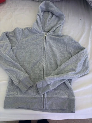 Ljusgrå zip hoodie med huva - En basic ljusgrå juicy couture hoodie med dragkedja framtill och huva. Tillverkad i mjukt sweatshirtmaterial, har långa ärmar och ribbade muddar vid ärmslut och nederkant. Perfekt för chill dagar eller när du vill ha en enkel och clean look.