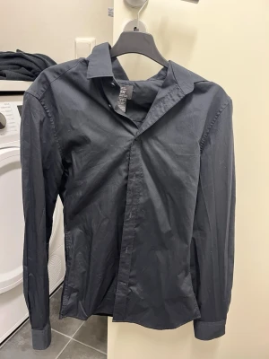 Svart skjorta med klassisk krage - Stilren svart skjorta med klassisk krage och långa ärmar. Skjortan har knappar framtill och är tillverkad i ett slätt bomullstyg. Perfekt för en clean och enkel look.