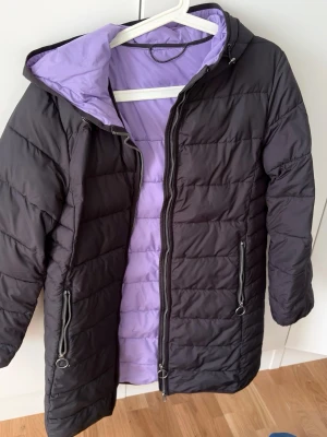 Svart lång dunjacka med lila foder - Säljer en svart lång höst jacka med lila foder och huva. Jackan har två dragkedjeförsedda fickor framtill och quiltad design. Perfekt för kalla vinterdagar och har en modern, stilren look.
