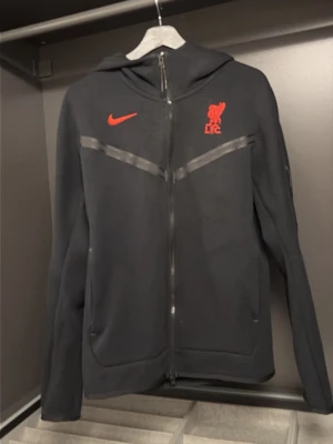 Svart Nike Liverpool hoodie - Svart hoodie från Nike med Liverpool FC-logga och Nike Swoosh i rött på bröstet. Hel dragkedja framtill, huva och diskreta fickor. Sportig design med tunna grå detaljer över bröstet. Perfekt för dig som älskar fotboll och vill ha en stilren look. Har lite knott på sig men änkelt att fixa.
