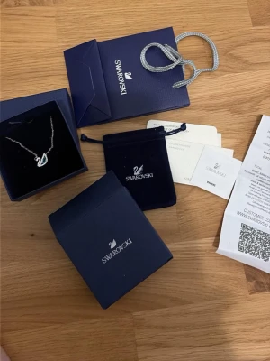 Swarovski svanhalsband med kristaller - Elegant halsband från Swarovski med ett silverfärgat kedja och ett hänge i form av en svan dekorerad med gnistrande vita och blå kristaller. Kommer i originalförpackning med sammetspåse och certifikat. Perfekt för dig som gillar stilrena smycken med lite extra bling.