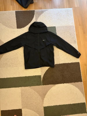 Svart vindjacka från Nike med huva - Snygg svart vindjacka från Nike med dragkedja framtill och praktisk huva. Jackan har två sidofickor och en diskret Nike-logga på bröstet. Tillverkad i lätt och vindtätt material, perfekt för blåsiga dagar.