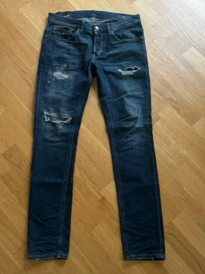 Dondup jeans geroge med hål - Säljer ett par mörkblå Dondup jeans i modellen George med slitningar och hål på både lår och knä. Jeansen har en smal passform och klassisk femficksdesign. 