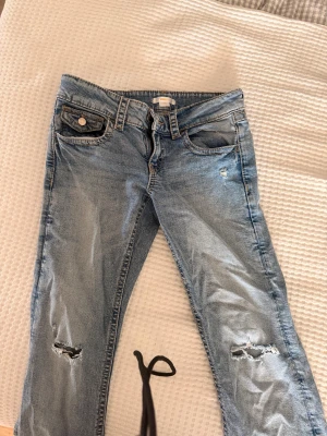 Ljusblå slitna jeans Gina Tricot - Säljer ett par ljusblå jeans från Gina Tricot i storlek 158. Jeansen har slitningar vid knäna och en cool tvättad look. Klassisk femficksmodell med knapp och dragkedja. Perfekta för en avslappnad och trendig stil.