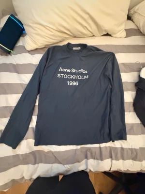 Mörkblå långärmad topp Acne Studios - Säljer en mörkblå långärmad topp från Acne Studios med trycket 'Acne Studios STOCKHOLM 1996' på bröstet. Klassisk passform och rund halsringning. Perfekt för dig som gillar stilrena och tidlösa plagg.