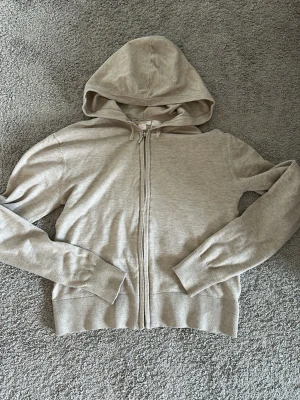 Beige zip-hoodie med huva - Ljusbeige hoodie med dragkedja framtill och justerbar huva. Tillverkad i mjukt, tunt material med ribbade muddar vid ärmslut och nederkant. Perfekt för en avslappnad look och enkel att matcha med andra plagg.