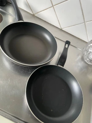 Två svarta stekpannor - Säljer två svarta stekpannor i non-stick material med plast- och gummihandtag. Perfekta för att laga allt från pannkakor till ägg. Båda har rund form och hål i handtaget för enkel upphängning. Passar till alla typer av kök.