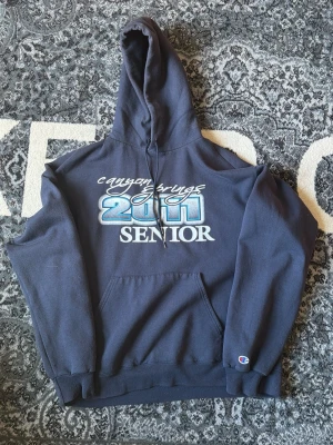 Mörkblå Hoodie med tryck från Champion - Mörkblå hoodie med stor huva och magficka. Framtill står det 'Canyon Springs 2011 Senior' i vitt och blått tryck. Baksidan har ett stort citat i vitt och grått. Klassisk Champion-logga på ärmen. Perfekt för dig som gillar retro college-stil.