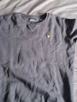 Grå sweatshirt från Lyle & Scott - Snygg grå sweatshirt från Lyle & Scott med klassisk rund halsringning och deras ikoniska gula logga broderad på bröstet. Tröjan har ribbade muddar vid ärmslut och hals. Perfekt för en avslappnad och stilren look.