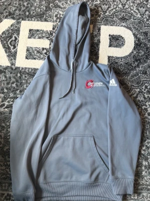 Ljusgrå Hoodie Cyclones Adidas - Ljusgrå hoodie med Cyclones Basketball-tryck på bröstet och Adidas-logga på ärmen. Klassisk känguruficka framtill, ribbade muddar och dragsko i huvan. Mjuk bomullskänsla och avslappnad passform, perfekt för chill dagar.