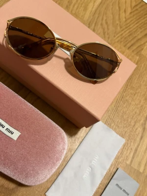 Runda solglasögon från Miu Miu - Snygga runda solglasögon från Miu Miu med guldfärgade metallbågar och bruna glas. Unik detalj på skalmarna och klassisk logga på sidan. Kommer med originalfodral och putsduk. Perfekta för dig som vill sticka ut med en trendig accessoar.