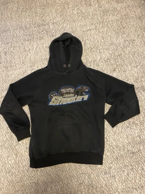 Svart Trapstar hoodie med tryck - Svart hoodie från Trapstar med stort tryck på bröstet där det står 'Trapstar London Shooters' och grafik av två pantrar. Klassisk känguruficka framtill och huva med dragsko. Tillverkad i mjukt material, perfekt för en avslappnad streetwear-look.