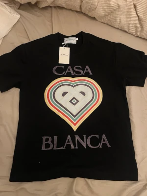 Svart t-shirt från Casablanca - Svart t-shirt från Casablanca med stort färgglatt hjärtmotiv och texten 'Casa Blanca' på framsidan. T-shirten har rund hals och korta ärmar. Materialet ser ut att vara mjuk bomull, perfekt för en trendig och avslappnad look. Den sitter även som mellan M och S