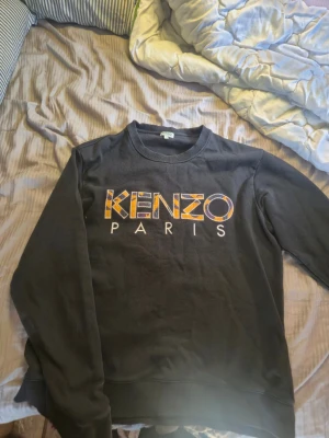 Svart Kenzo Paris sweatshirt - Säljer en svart sweatshirt från Kenzo med stort broderat Kenzo Paris-tryck i gult, lila och vitt på bröstet. Tröjan har rund halsringning och ribbade muddar vid ärmslut och nederkant. Perfekt för dig som gillar streetwear och vill sticka ut. Har storlek m men dom är små i storlek så fick ta l
