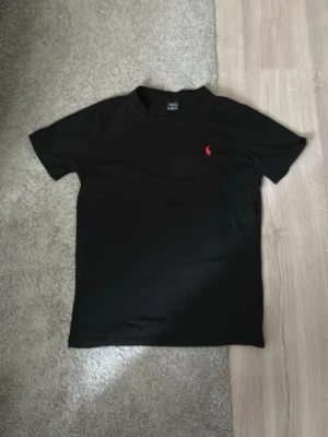 Svart t-shirt med röd logga - Snygg svart t-shirt med klassisk röd broderad logga på bröstet. T-shirten har korta ärmar och rund halsringning. Tillverkad i mjuk bomull som känns skön mot huden. Perfekt till jeans eller shorts för en clean och enkel look.