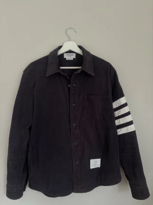 Thom Browne overshirt i 100 % ull-cord, laget i Japan med Cross Company-label. - Sjelden arkivmodell fra den tidlige japanske produksjonen, sannsynligvis fra rundt 2008–2012. Det som gjør denne spesielt interessant er stoffet – 100 % ull-cord i stedet for vanlig bomulls-cord. Kraftig stoff med en veldig fin tekstur.  Mørkegrå/charcoal med 4-Bar på ermet, gullfargede trykknapper, hvitt pipingarbeid på innsiden og cupro-fôr.  Dette er et plagg laget for å vare. Kraftig ullstoff, solid konstruksjon og gode materialer gjør at dette fint kan brukes i mange år fremover. Med riktig