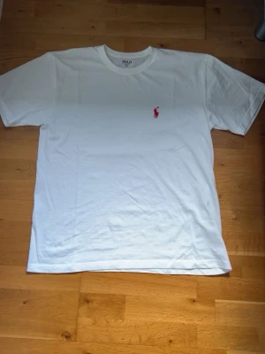 Vit t-shirt från Polo Ralph Lauren - Klassisk vit t-shirt från Polo Ralph Lauren med röd broderad logga på bröstet. T-shirten har rund halsringning och korta ärmar. Tillverkad i mjuk bomull för en skön känsla och enkel stil.