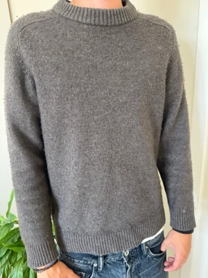 Mörkgrå stickad tröja - Mysig mörkgrå stickad tröja med rund halsringning och ribbade muddar vid ärmslut och nederkant. Tröjan har en enkel och stilren design som passar perfekt till jeans. Materialet ser ut att vara ull eller en ullblandning. Syns inte men mer av en grå brun färg 