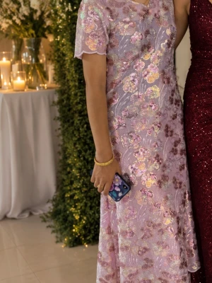 Ljuslila blommig långklänning - Elegant långklänning i ljuslila med glittriga blommiga broderier. Klänningen har korta ärmar och v-ringning, perfekt för speciella tillfällen. Materialet har en skimrande finish och fina spetsdetaljer vid ärmslut och halsringning. Storlek xs 