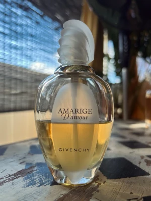 Givenchy Amarige D'amour  50ml flaska;vintage - Säljer en Amarige D'amour parfym från Givenchy. Flaskan är elegant och genomskinlig med en mjukt gul vätska och ett vitt, snirkligt lock. Designen är lyxig och tidlös, perfekt för dig som gillar exklusiva dofter