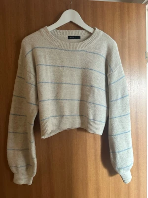 Beige cropped stickad tröja SHEIN - Säljer en beige cropped stickad tröja från SHEIN med tunna ljusblå ränder. Tröjan har rund halsringning, långa ärmar och ribbade muddar. 