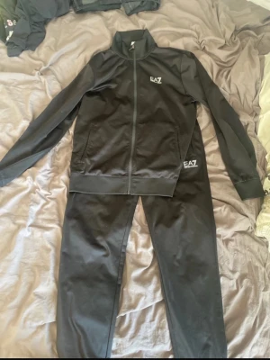 Svarta mjukisdress från EA7 - Säljer ett par svarta mjukisbyxor och zip hoodie från EA7 med logga på vänster ben och bröst. Byxorna har resår i midjan och sidofickor.  Perfekta för chill eller träning, tillverkade i ett mjukt och bekvämt material.