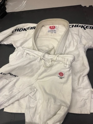 Vit BJJ gi från Chokem, storlek A1 - Säljer en vit brasiliansk jiu-jitsu gi från Chokem i storlek A1. Dräkten består av jacka och byxor med broderade loggor på ärm och ben. Materialet är slitstarkt och tjockt, perfekt för träning och tävling. Klassisk design med förstärkta sömmar och dragsko i midjan.