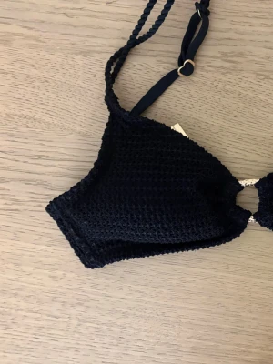 Svart bikiniöverdel med struktur - Helt ny, endast provad och tvättad en gång. Nypris ca 500kr. Säljer en svart bikiniöverdel med snygg struktur och dekorativ ringdetalj framtill. Modellen har smala axelband och trekantsformade kupor. Perfekt för strandhäng eller poolparty!