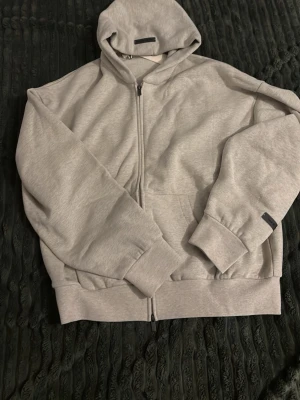 Grå Essentials Fear of God hoodie - Snygg grå zip hoodie från Essentials Fear of God med huva och fickor framtill. Stort tryck på ryggen med 'ESSENTIALS FEAR OF GOD'. Tillverkad i mjukt sweatshirtmaterial, perfekt för en avslappnad streetwear-look.