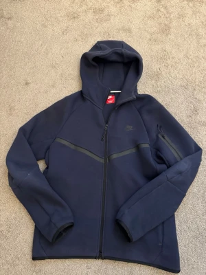 Mörkblå Nike hoodie med dragkedja - Snygg mörkblå hoodie från Nike med hel dragkedja framtill och huva. Hoodien har diskreta svarta detaljer över bröstet och en ficka med dragkedja på ena ärmen. Tillverkad i ett mjukt och tjockt material som passar perfekt för kyligare dagar.