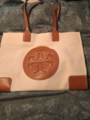Beige tygväska med bruna detaljer - Stilren handväska i beige tyg med bruna läderdetaljer och stora handtag. Väskan har ett stort brunt emblem framtill och förstärkta hörn i samma material. Perfekt för dig som gillar klassisk och enkel design.. ej äkta . Aldrig använt.