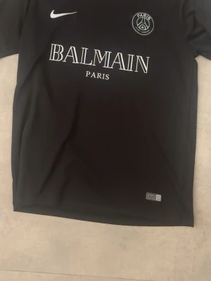 Svart PSG x Balmain matchtröja Nike - Snygg svart matchtröja från Nike i samarbete med PSG och Balmain. Tröjan har rund hals, vita tryck med Paris Saint-Germain-logga och Balmain Paris på bröstet. Tillverkad i Dri-FIT material som andas, perfekt för träning eller streetwear.
