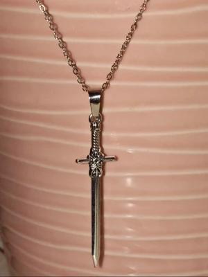 Halsband med svärdhänge i silverfärg - Snyggt halsband med ett detaljerat svärdhänge i silverfärgad metall. Kedjan är tunn och smidig, och svärdet har en cool, stilren design som passar perfekt till streetwear eller för dig som gillar unika accessoarer.