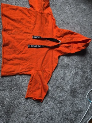 Orange hoodie med tryck på snöre (maghoodie) - Säljer en orange hoodie med huva och svarta snören där det står 'TRICK ME ONCE CAN'T FOOL ME TWICE'. Hoodien har kortare passform och vida ärmar. Perfekt för dig som vill sticka ut med en färgstark och unik stil.