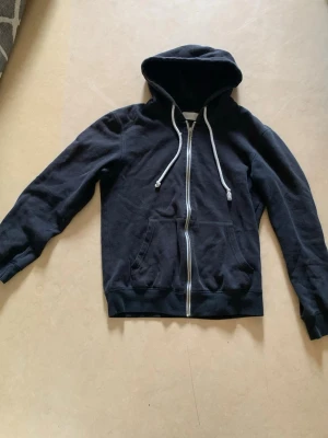 Svart zip hoodie med huva - Klassisk svart hoodie med dragkedja framtill och vita dragsnören i huvan. Hoodien har två fickor på magen och ribbade muddar vid ärmslut och nederkant. Perfekt för en avslappnad och stilren look.