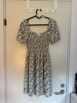 Blommig midiklänning från H&M - Söt blommig midiklänning från H&M med puffärmar och smockad överdel. Klänningen har svart, vit och gul blommönster, fyrkantig ringning och är gjord i ett mjukt, stretchigt material. Perfekt för en trendig och avslappnad look. Klänningen har bara andväns 1 gång så den är nästan som ny. 