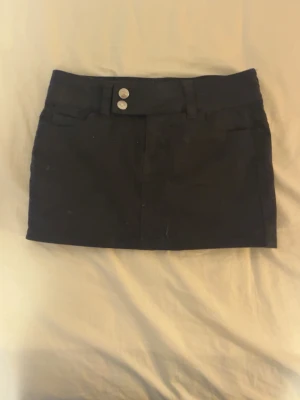 Svart kortkjol med knappar - Stilren svart kortkjol med två dekorativa silverknappar i midjan och bred linning. Kjolen har en klassisk rak passform och är enkel att matcha till olika outfits. Perfekt för dig som gillar en clean och tidlös look.