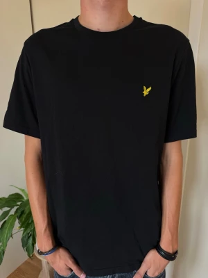 Svart t-shirt med gul logga - Svart t-shirt med rund hals och en liten gul broderad logga på bröstet. Klassisk passform och korta ärmar, perfekt till jeans eller shorts. Tillverkad i mjuk bomull för en skön känsla hela dagen.