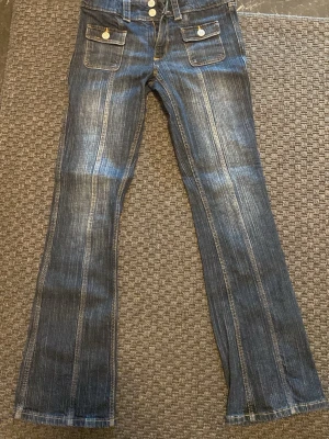 Mörkblå bootcut jeans med fickor - Säljer ett par mörkblå bootcut jeans med markerade sömmar och två stora fickor framtill med knappar. Jeansen har utsvängda ben och dubbla knappar i midjan. 