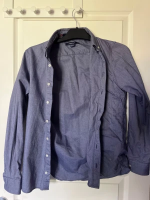 Blå skjorta från Gant - Klassisk blå skjorta från Gant med lång ärm och knappar framtill. Skjortan har en tidlös design med button-down krage och är tillverkad i mjuk bomull. Perfekt för en clean och stilren look.