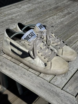 Diesel vita sneakers - Snygga vita sneakers från Diesel med svart logodetalj på sidan och gråa skosnören. Skorna har låg profil och klassisk design med rund tå och platt sula. Materialet är läder och textil, perfekt för en cool och avslappnad look. Storlek 43 EU. Pris är inte hugget i sten. De ska se slitna ut som design. I övrigt fint skick men lite slitna inuti skorna på sulan. 