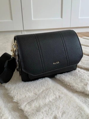 Svart axelväska från NA-KD - Snygg svart axelväska från NA-KD i fejkläder med guldfärgade detaljer och bred axelrem. Väskan har coola ormmönstrade sidor och praktiska fack. Perfekt för dig som vill ha en trendig och stilren accessoar till din outfit💗