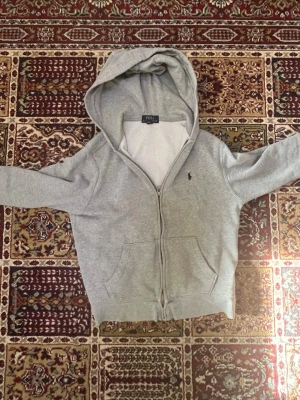 Grå zip hoodie från Polo Ralph Lauren - Snygg grå hoodie med dragkedja från Polo Ralph Lauren. Klassisk design med huva, två fickor framtill och den ikoniska lilla loggan broderad på bröstet. Tillverkad i mjukt sweatshirtmaterial som är perfekt för chill dagar.